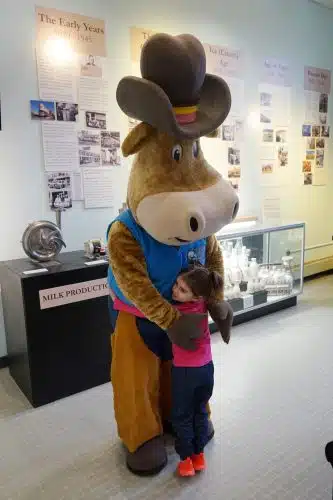 Byrne Dairy mascot Byrnsie, a brown cow in a blue vest and brown cowboy hat, hugs a child at the Byrne Dairy 90th anniversary exhibit at the MOST