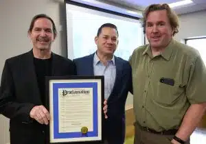 Three men smiling and holding a framed proclamation